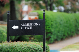 Closeup of sign "Admissions Office" with arrow, in front of a green hedge.